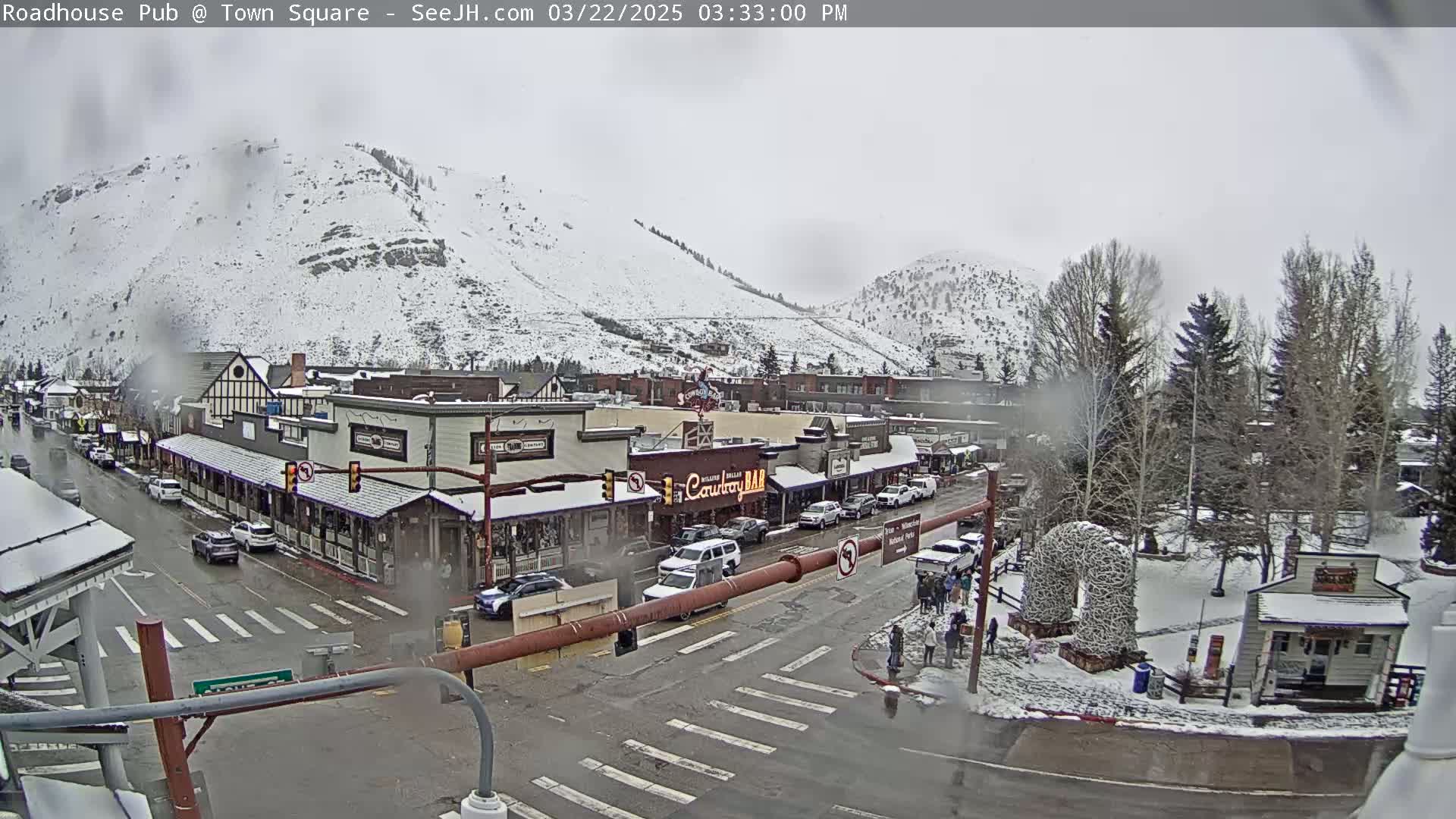 A snow-covered town square with cars parked along the street, surrounded by snow-dusted buildings and hills under an overcast sky.