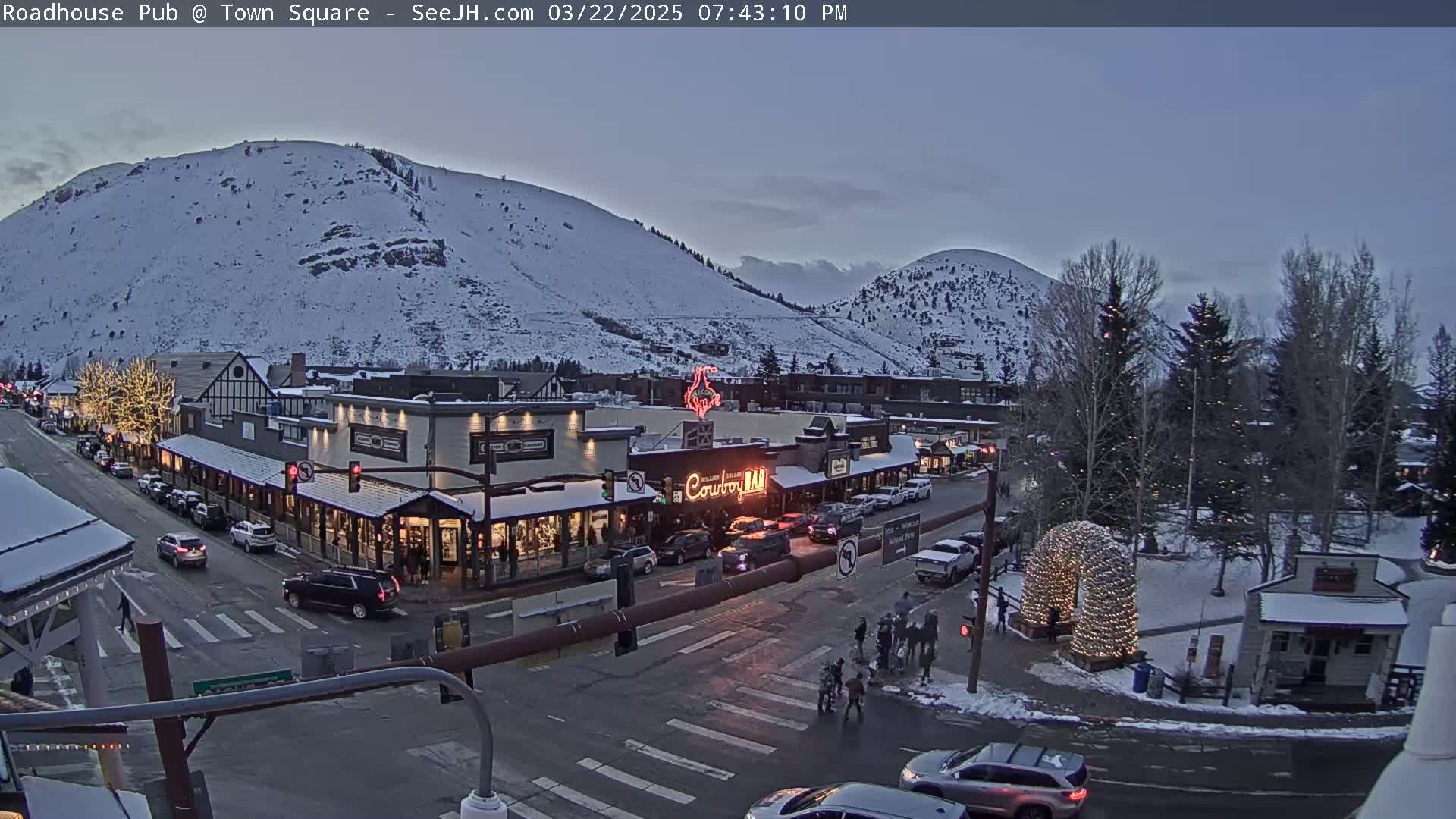 A snow-covered mountain overlooks a town square at dusk, where cars are parked along the streets and pedestrians are crossing the intersection near a lit-up archway.