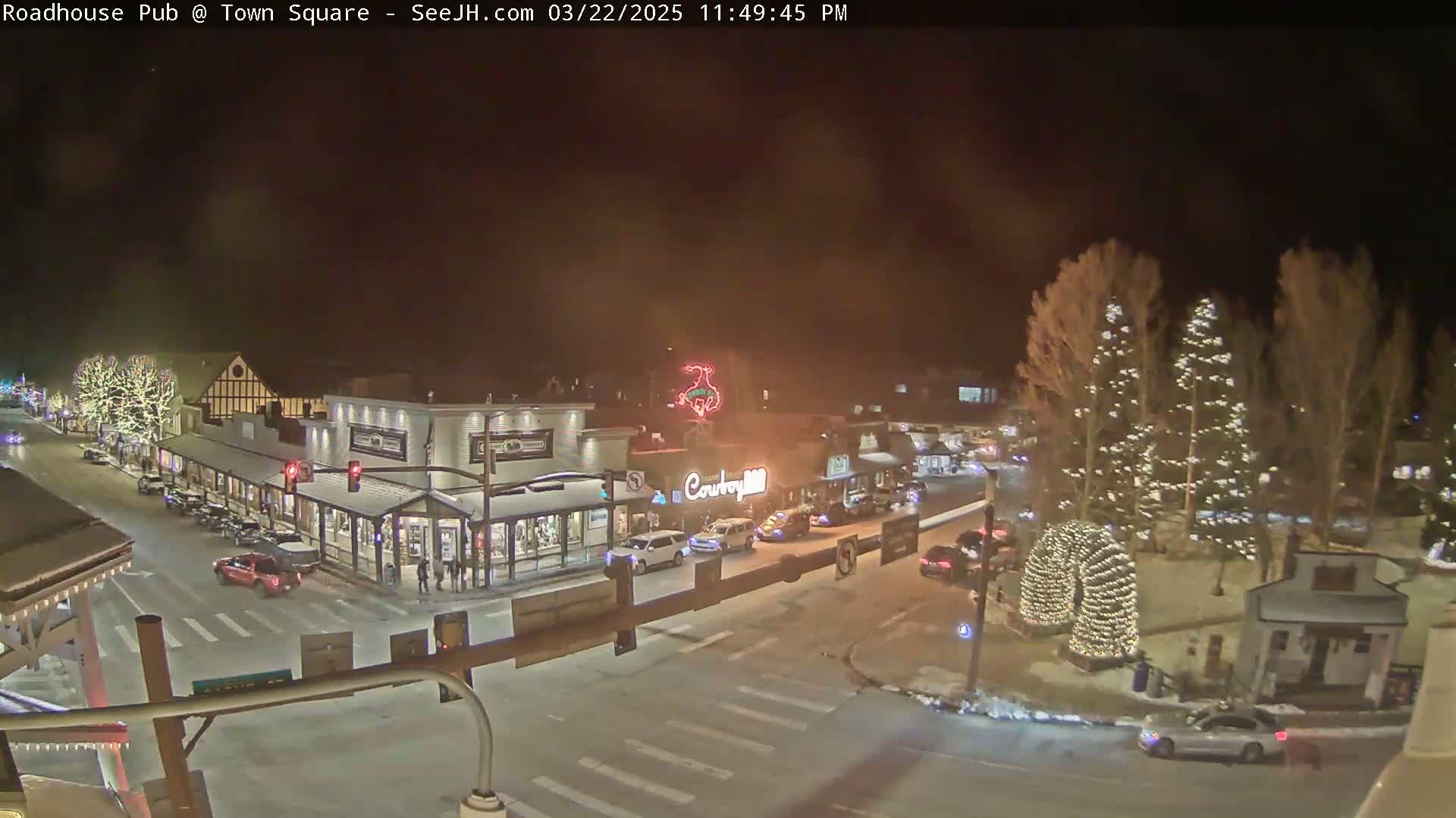 A nighttime view of a town square with several illuminated buildings, cars, and Christmas trees.