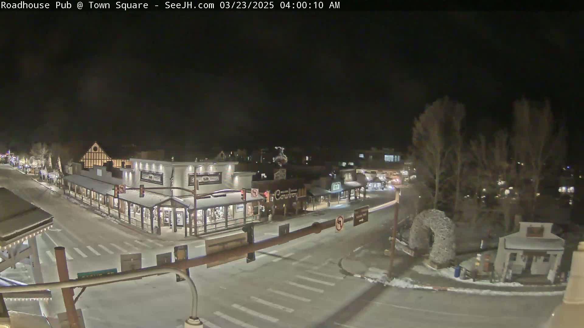 A snow-covered town square at night, viewed from above, shows several buildings with streetlights illuminating the intersection.