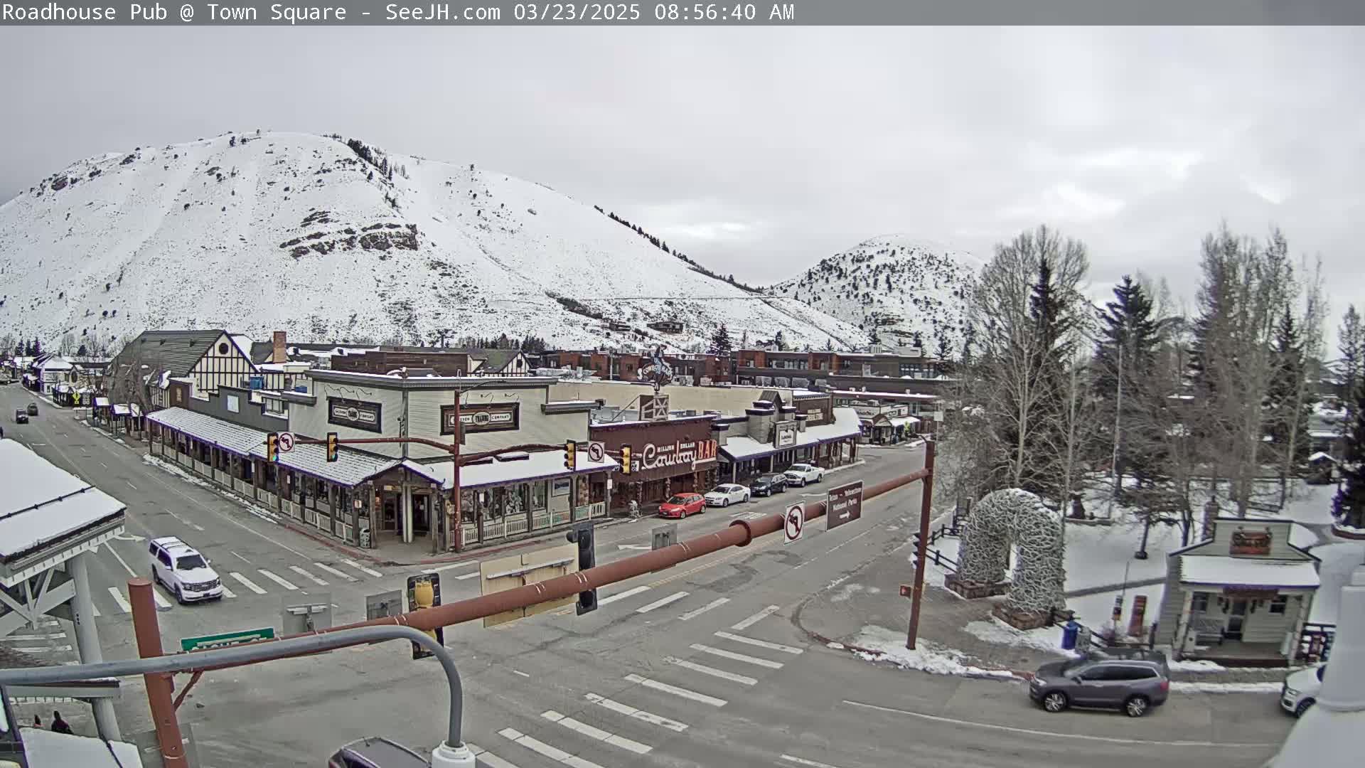 A snow-covered mountain overlooks a town square with several buildings, sparsely parked cars, and a few bare trees under an overcast sky.