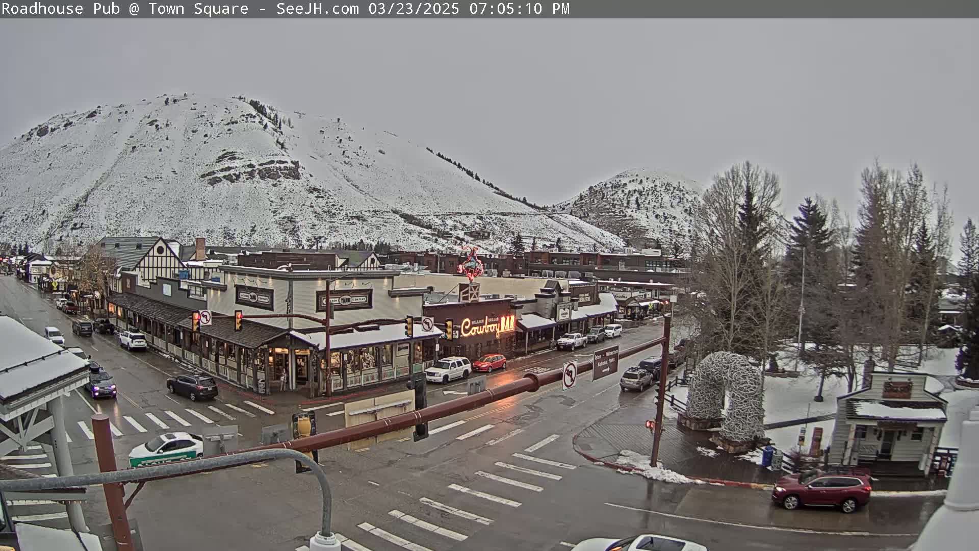 A snow-covered mountain overlooks a town square with several buildings, cars, and trees under an overcast sky.