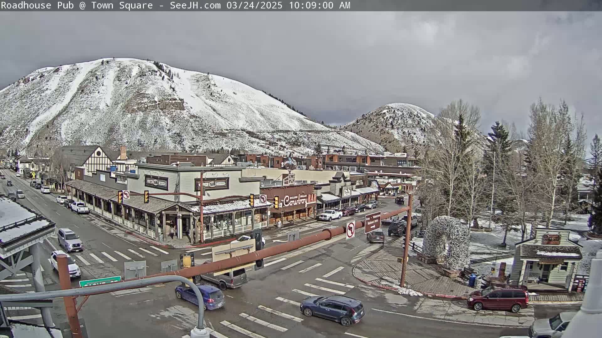 A snow-dusted mountainside overlooks a town square with several buildings and vehicles at an intersection on an overcast day.