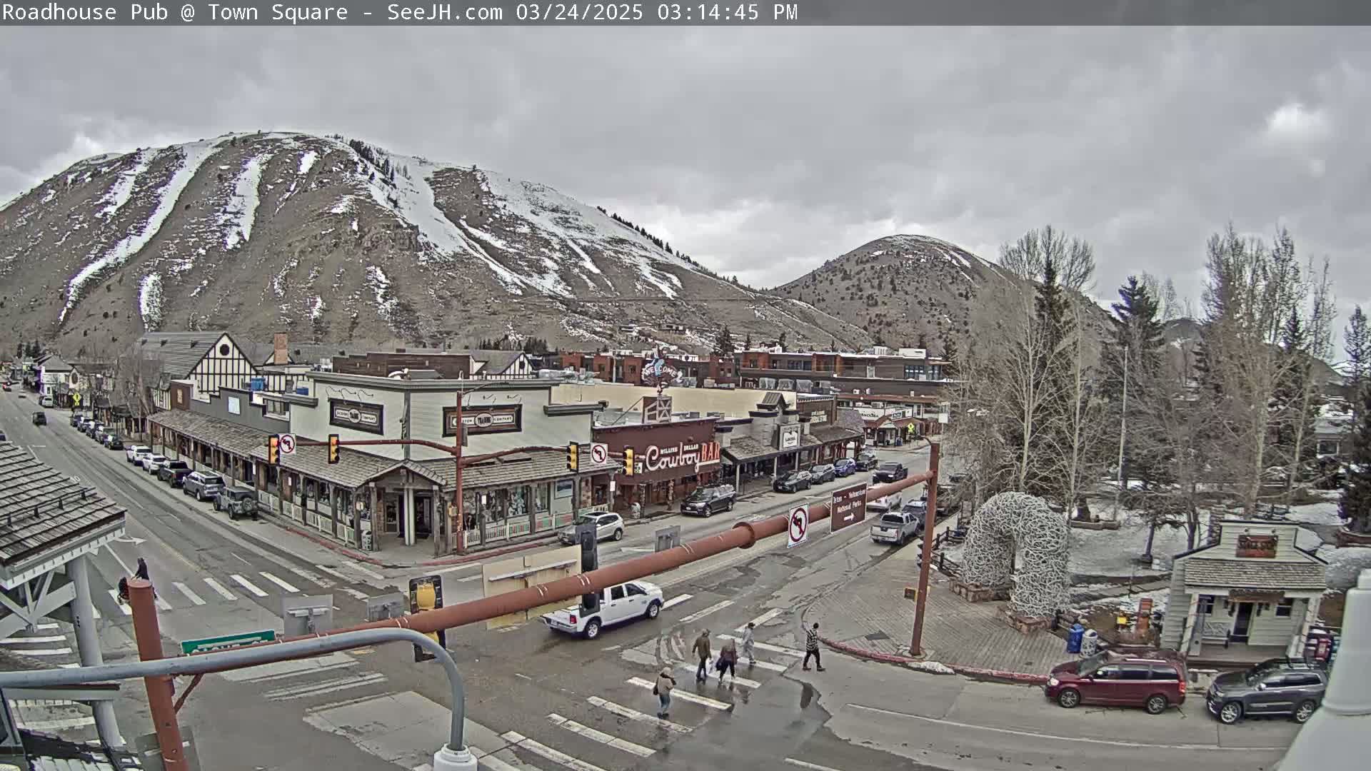 A town square with a few cars and pedestrians, situated at the base of a snow-dusted mountain under an overcast sky.