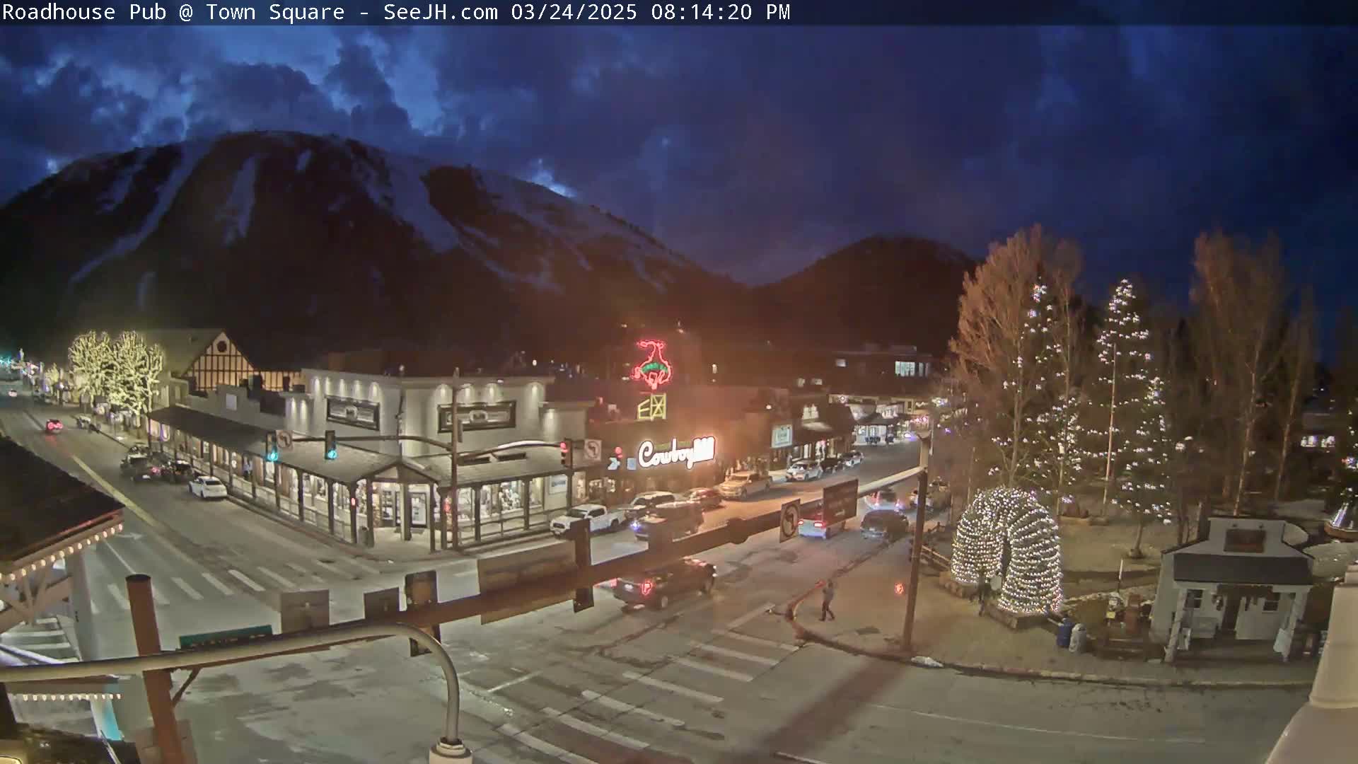 A nighttime view of a snow-covered mountain overlooking a town square with illuminated buildings and holiday lights under a partly cloudy sky.