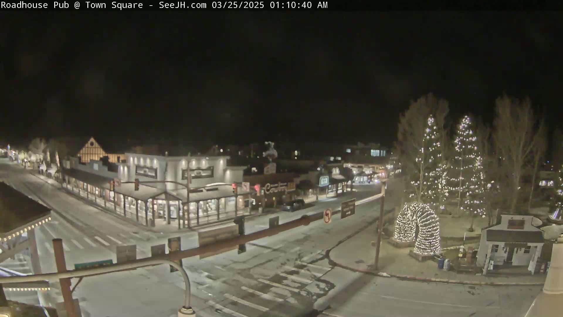 A snow-dusted town square at night, illuminated by streetlights and Christmas lights on trees.