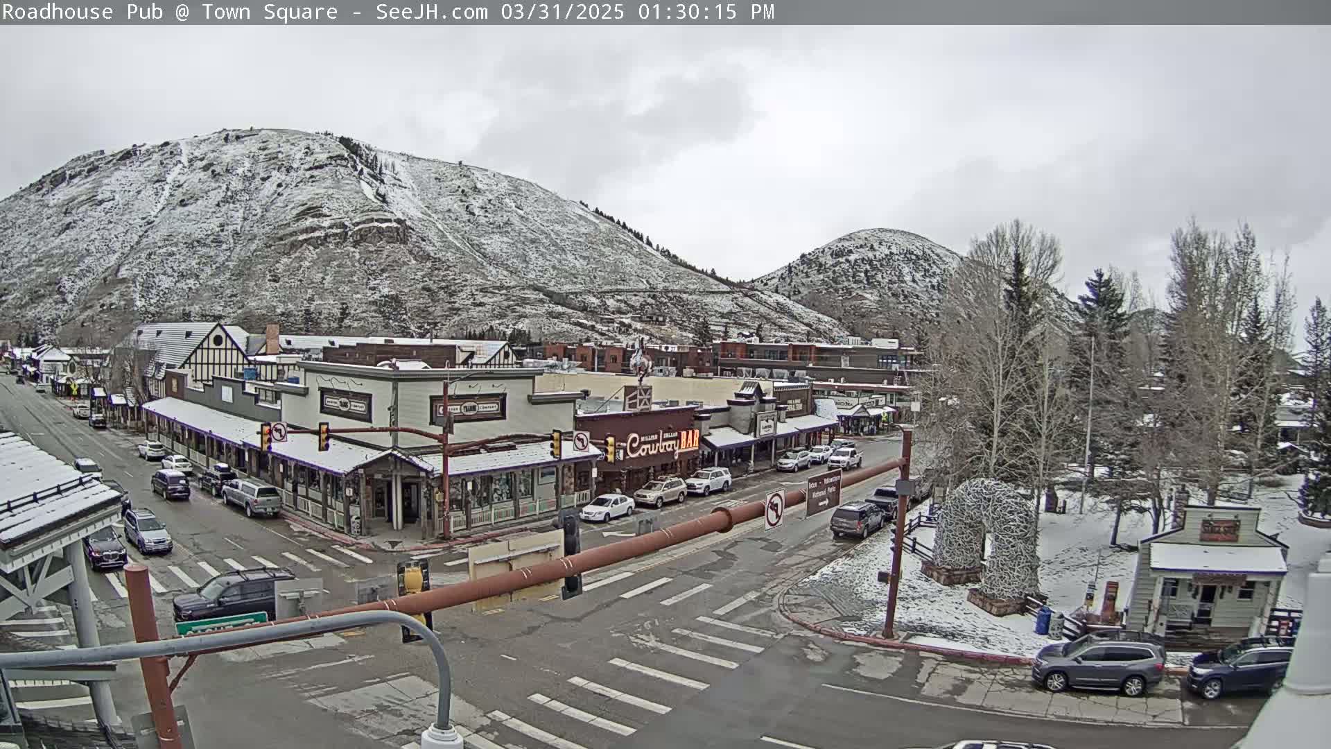 A snow-dusted mountainside overlooks a town square with cars parked along the streets, under an overcast sky.