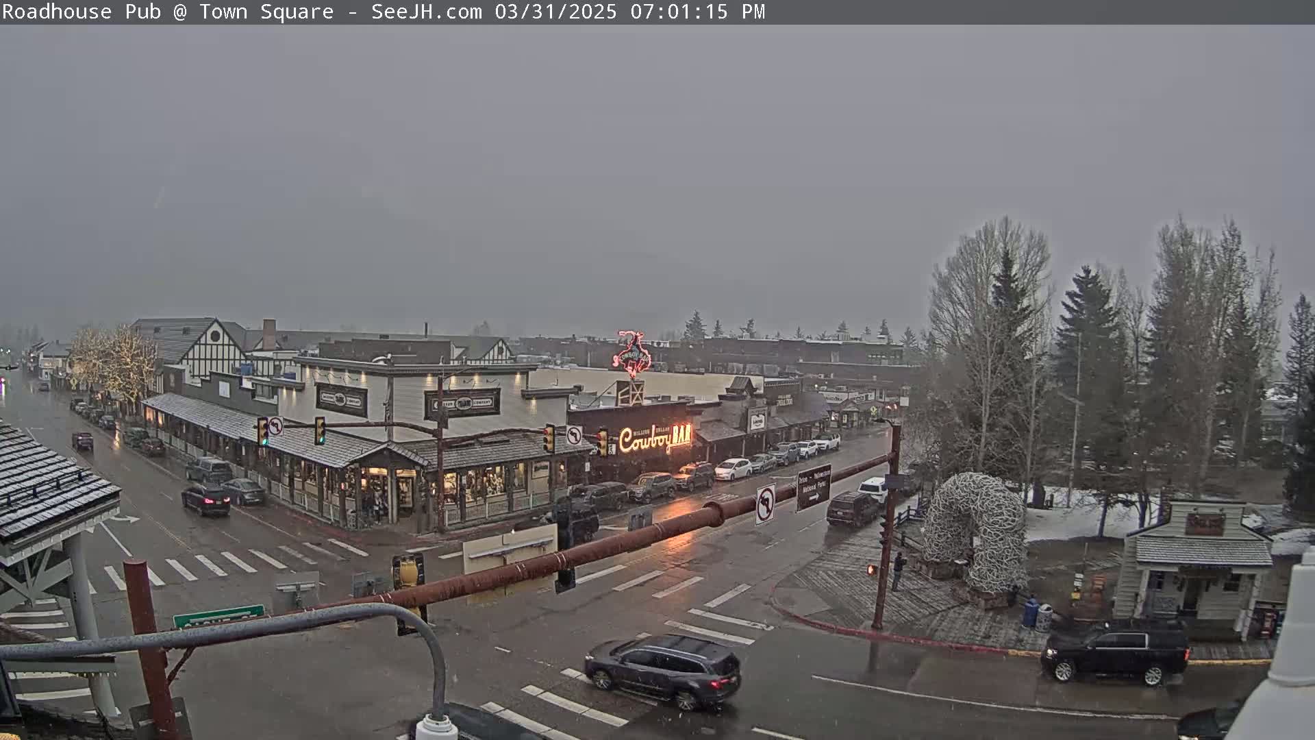 A snow-covered town square with several buildings, cars, and pedestrians, viewed from above on an overcast day.