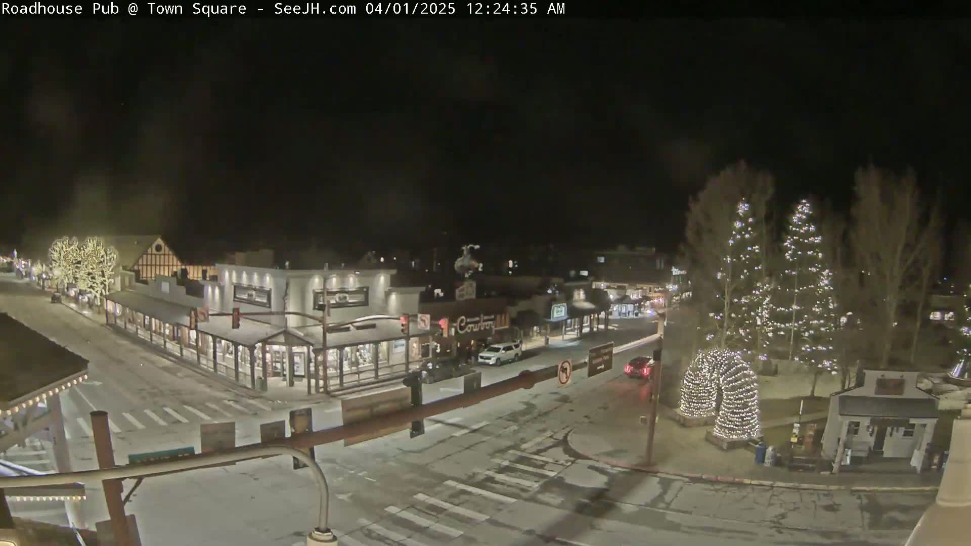 A snow-dusted town square at night, illuminated by streetlights and Christmas lights strung on trees and arches.