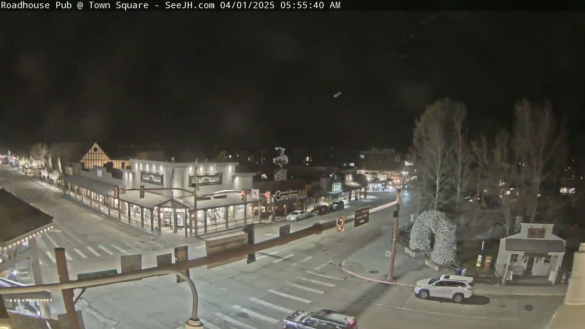 A nighttime view of a town square with several buildings, vehicles, and bare trees under a clear, dark sky.