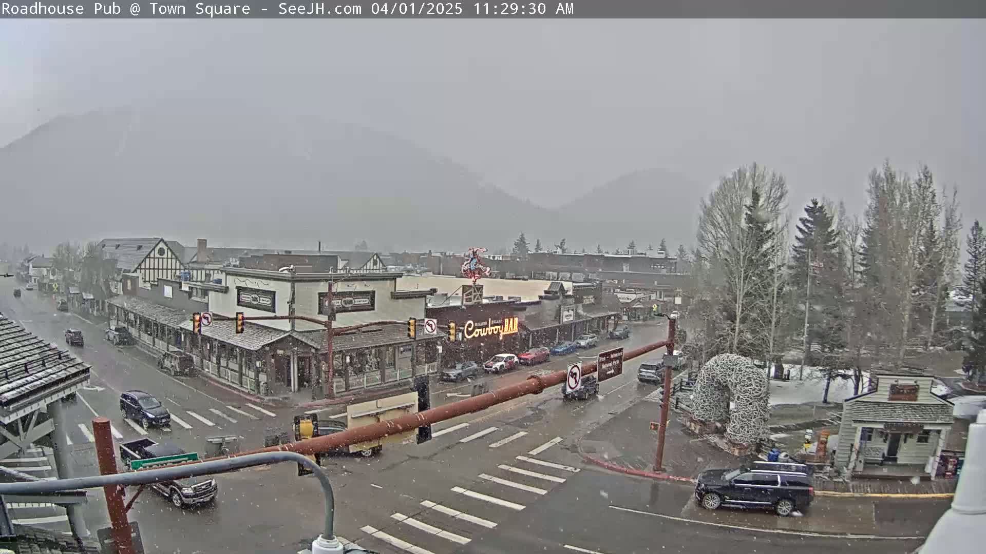 A snow-covered town square with cars and buildings is seen under a light snowfall, with mountains visible in the background.