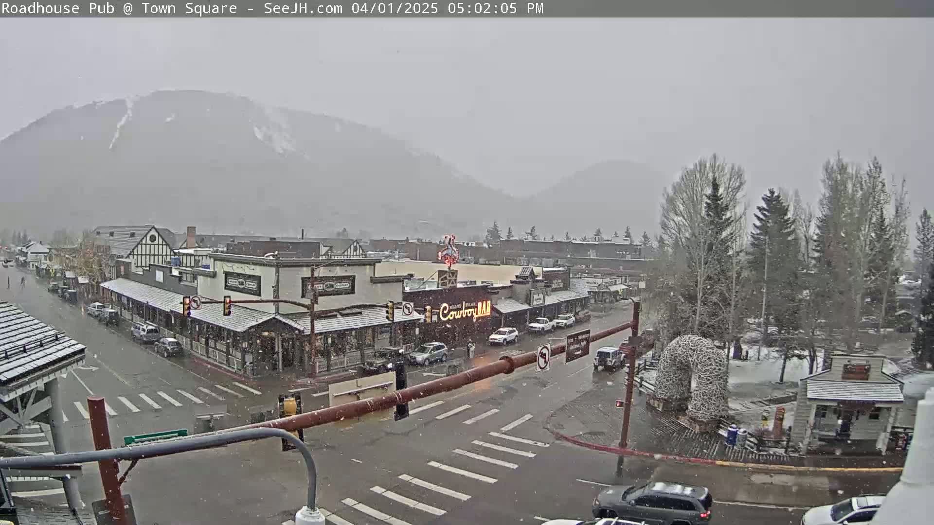 A snow-covered town square with buildings and cars, under a gray, snowy sky, with a mountain in the background.