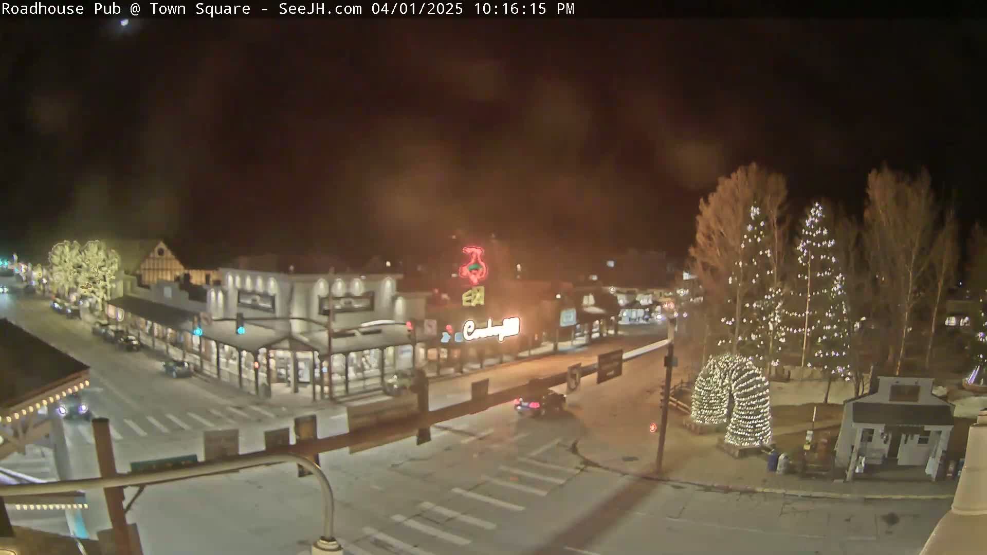 A nighttime, snow-covered town square with illuminated buildings and Christmas trees.