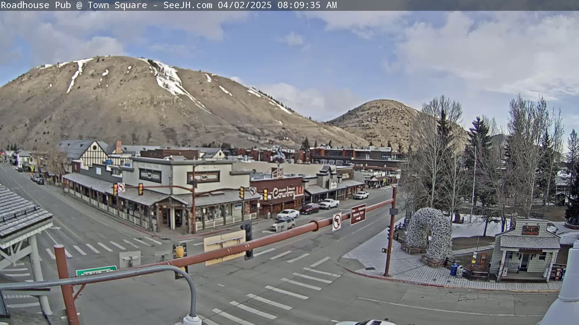 A mostly sunny day reveals a town square with several buildings, a few cars, and barren trees, all nestled at the base of two mountains with some remaining snow.