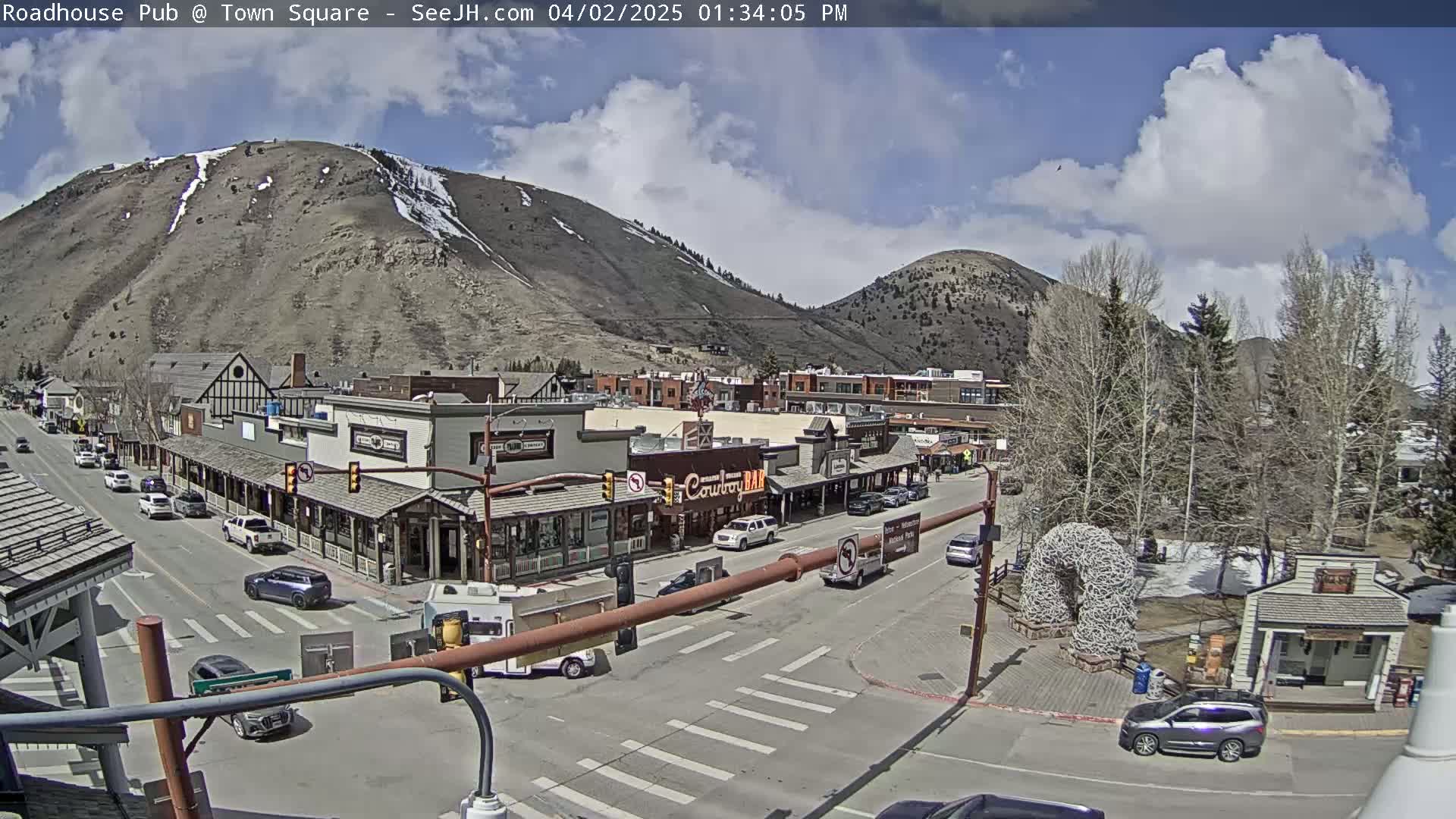 A partly sunny day reveals a mountain town square with several cars parked along the streets and buildings in the background, under a partly cloudy sky.