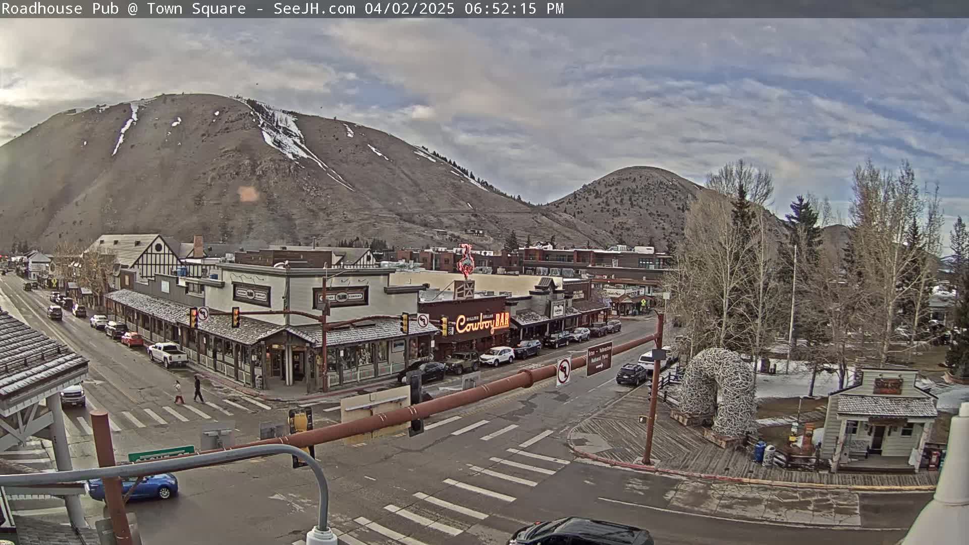 A partly cloudy day reveals a mountain town square with several buildings, cars parked along the streets, and a pedestrian crossing.