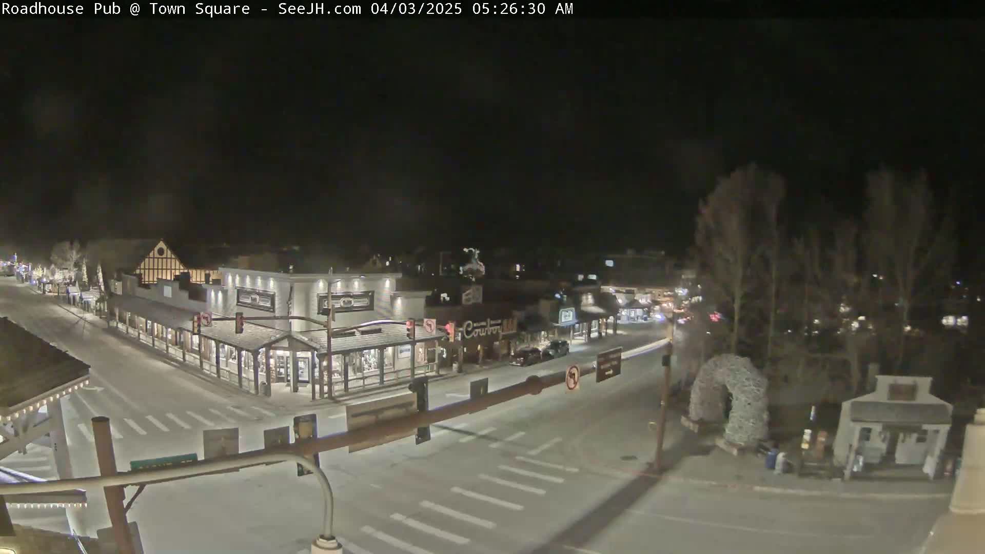A snow-covered town square at night, illuminated by streetlights, shows several buildings and a few cars.