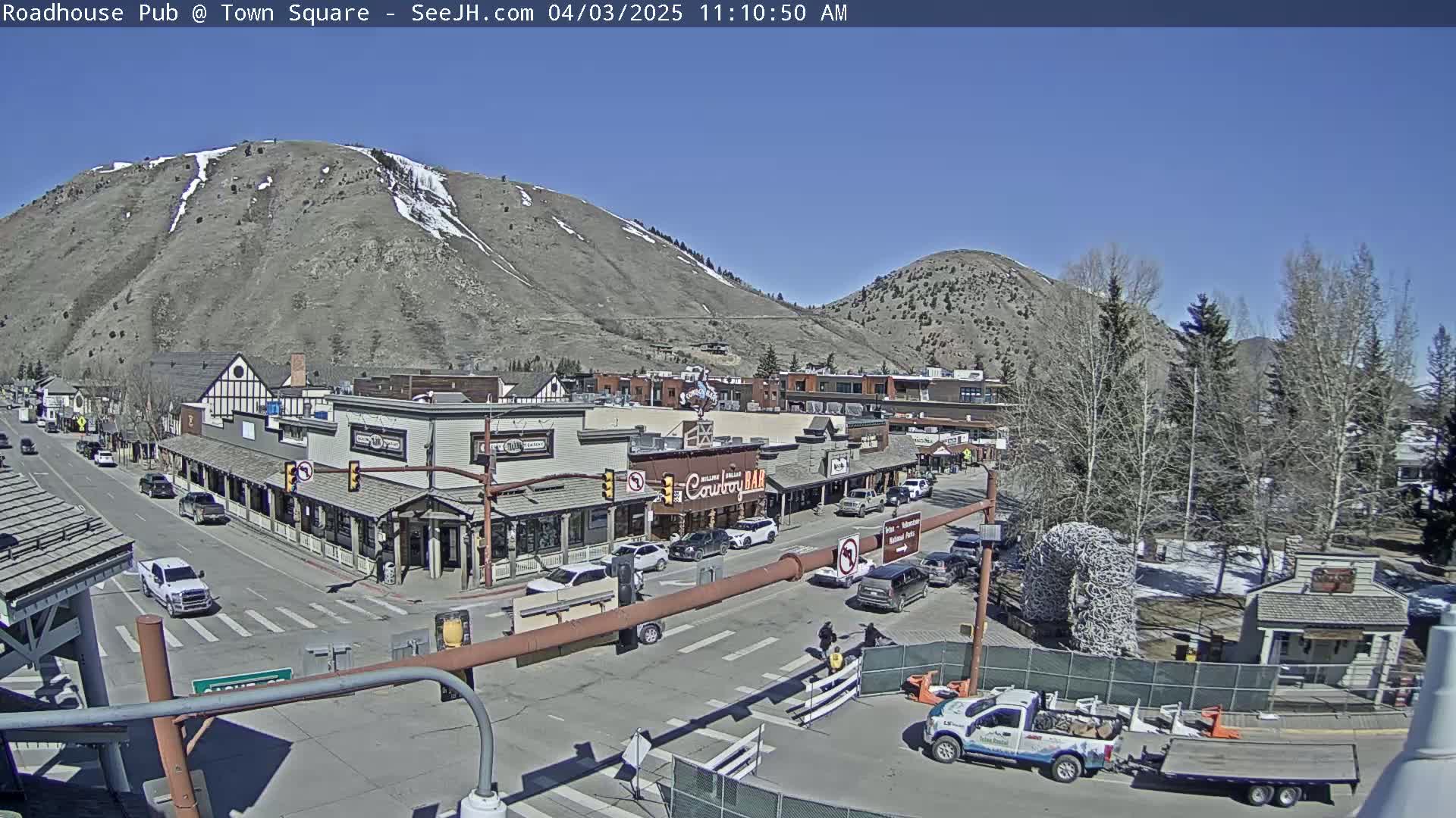 A sunny day reveals a town square nestled between mountains, with cars parked along the streets and buildings lining the square.