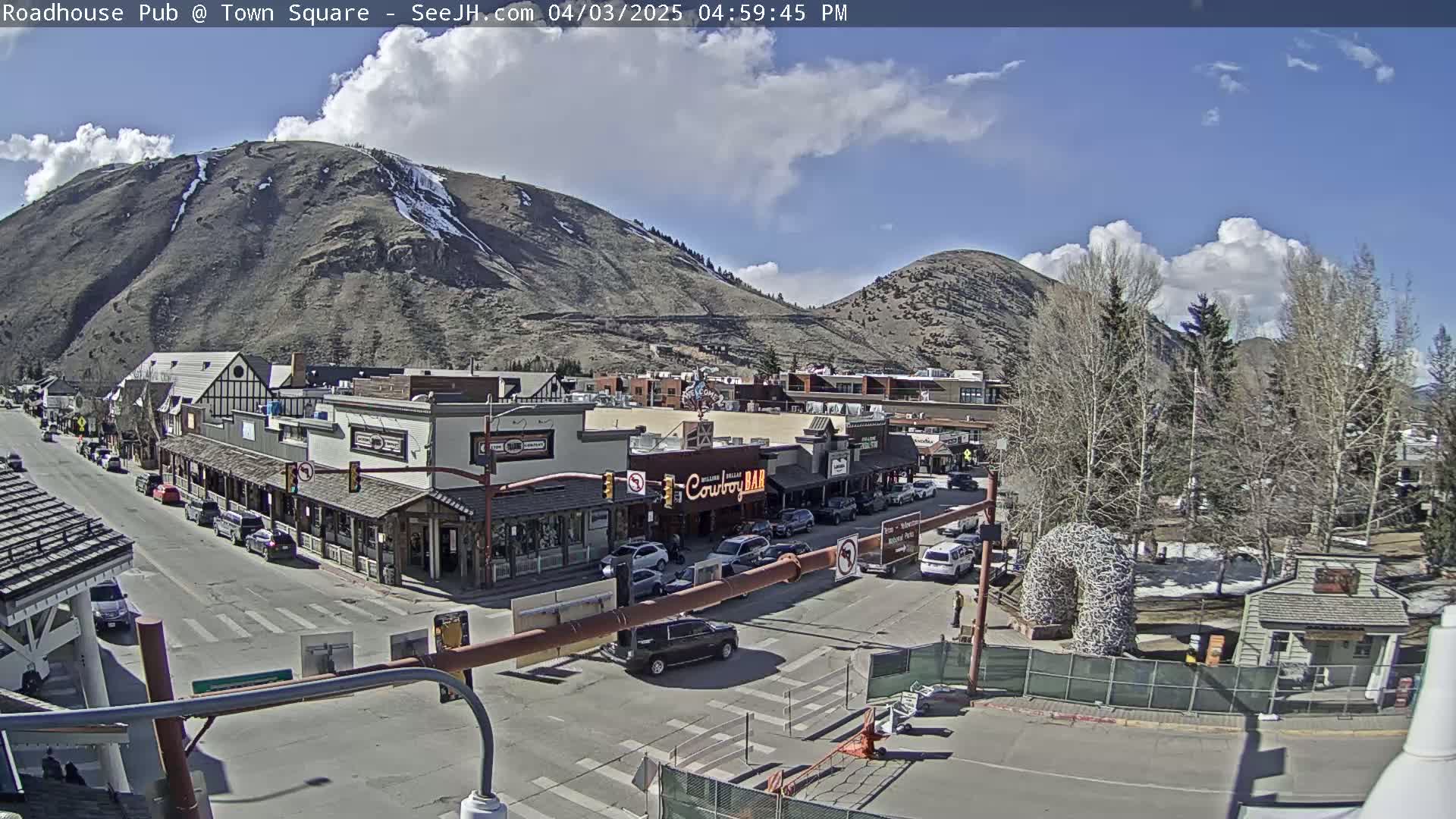 A partly sunny day reveals a mountain town square with buildings, cars, and trees, featuring a large, mountainous backdrop with patches of snow.