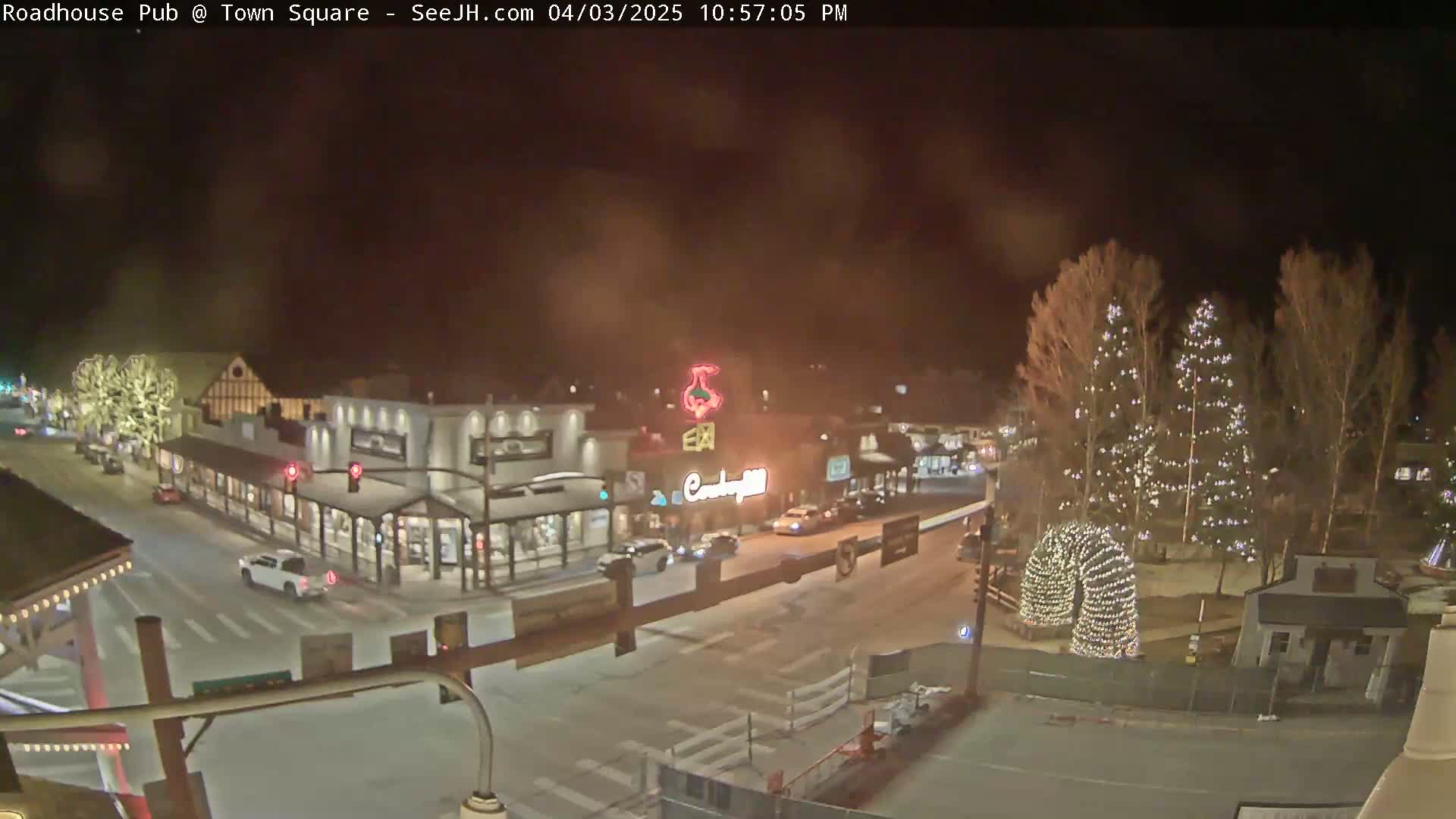 A nighttime view of a town square with illuminated buildings and Christmas trees, under a hazy sky.