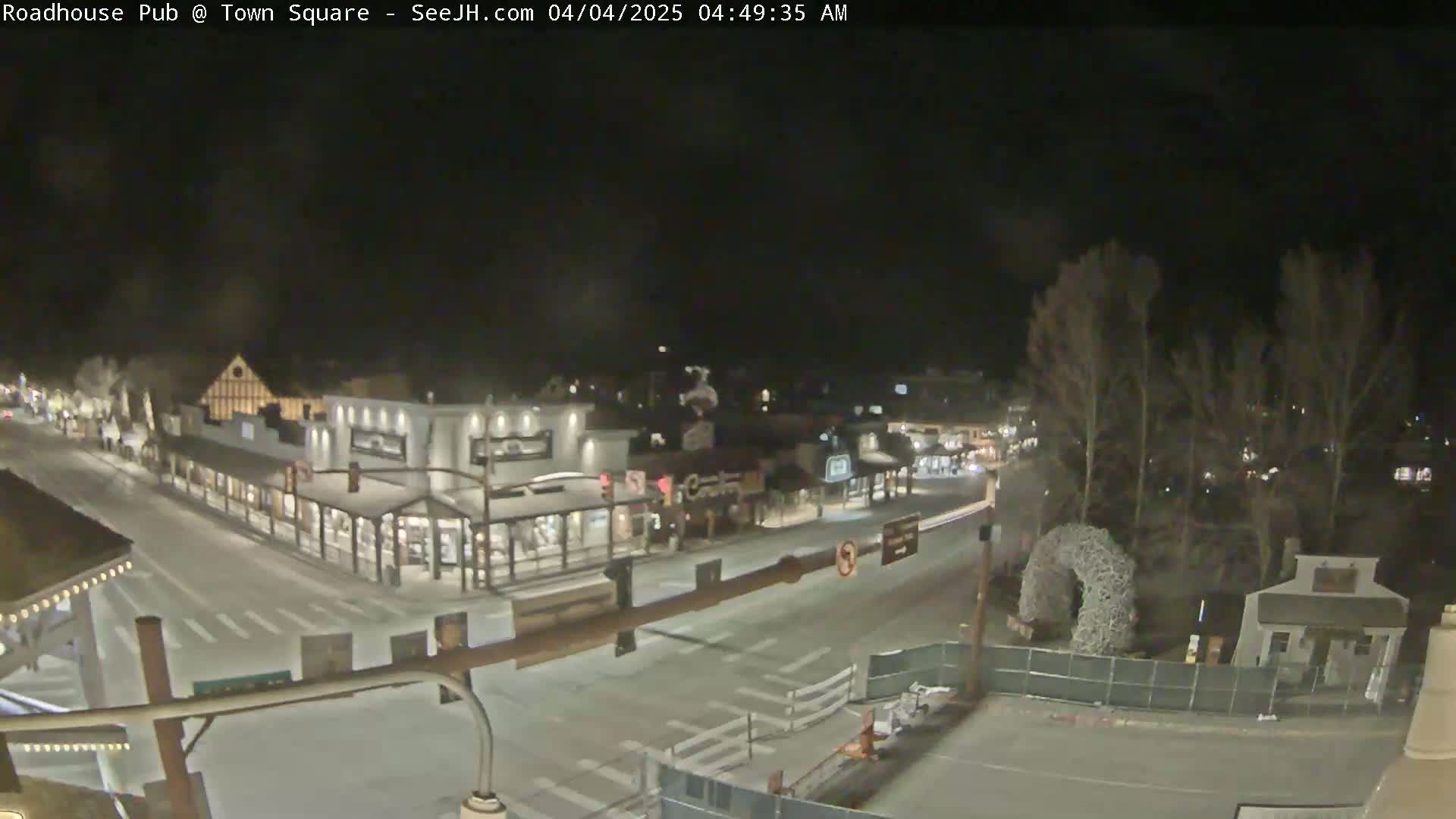 A nighttime aerial view of a snow-covered town square with several buildings and sparsely trafficked streets.