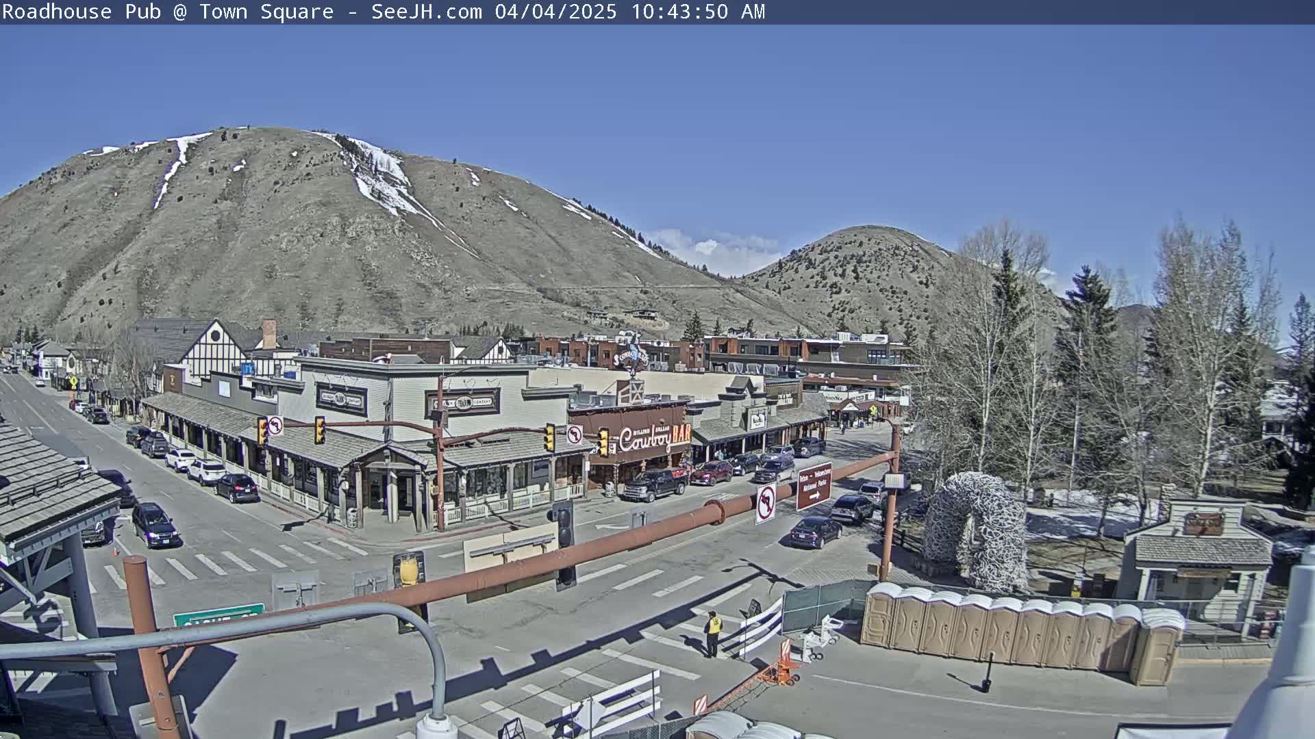 A sunny day in a mountain town shows a street scene with buildings, cars, and a large antler arch.