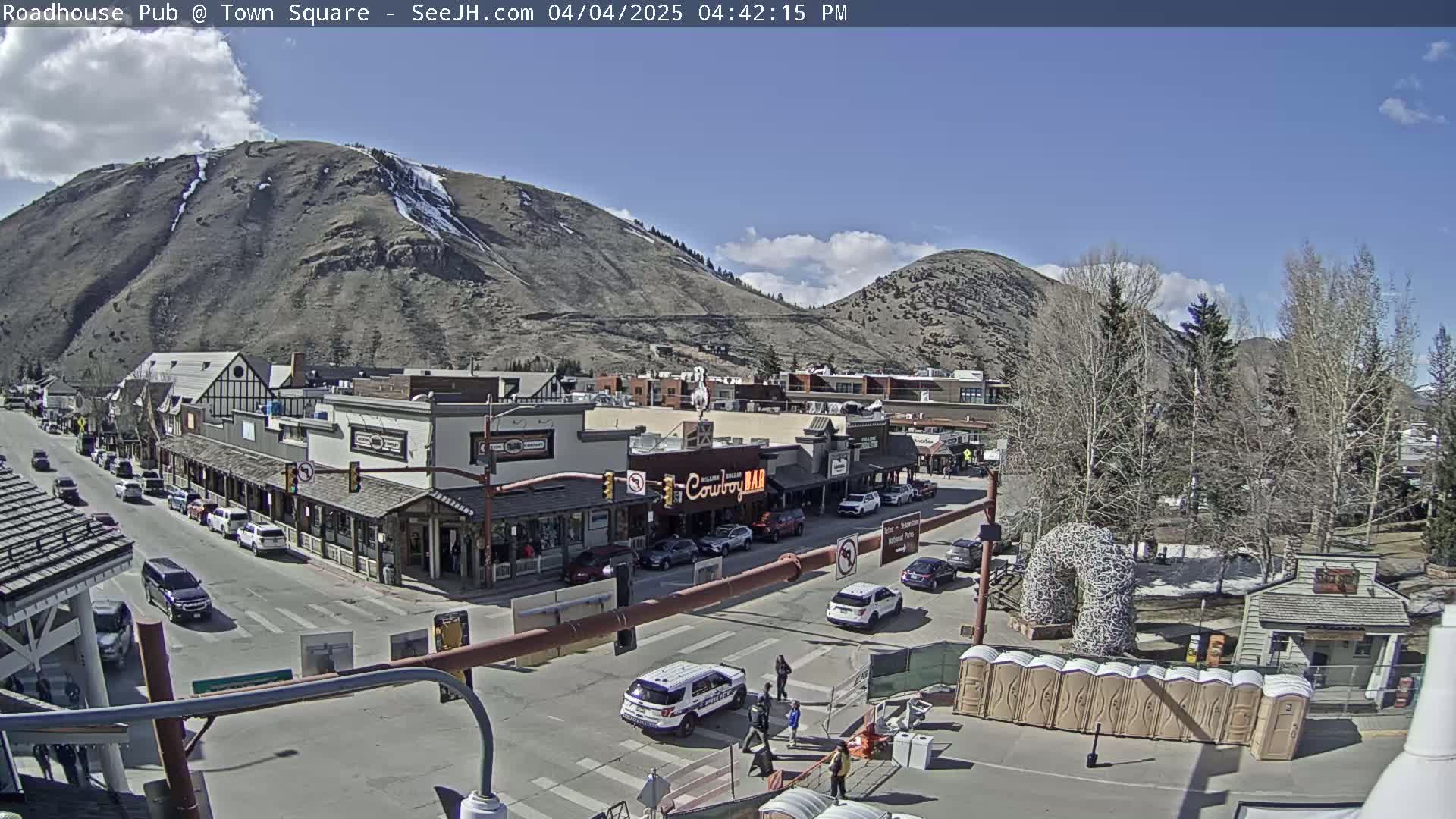 A partly sunny day reveals a mountain town with several buildings lining a street, with cars parked and driving, and a few pedestrians present.