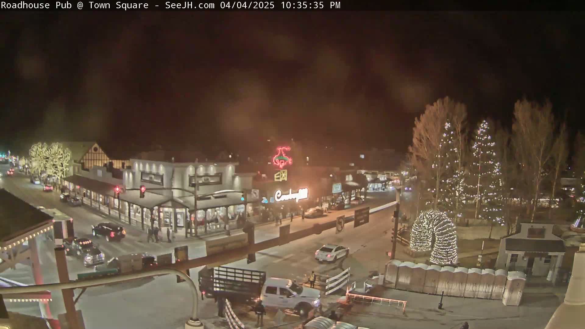 A nighttime view of a town square with snow-covered streets, illuminated buildings, and Christmas lights on trees.