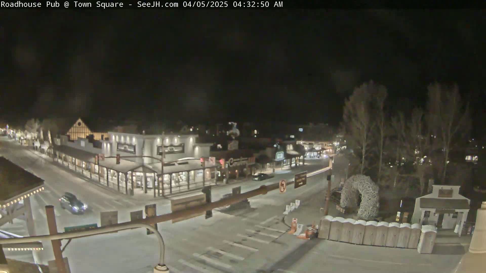 A snow-covered town square at night, illuminated by streetlights, shows several buildings and a few cars.