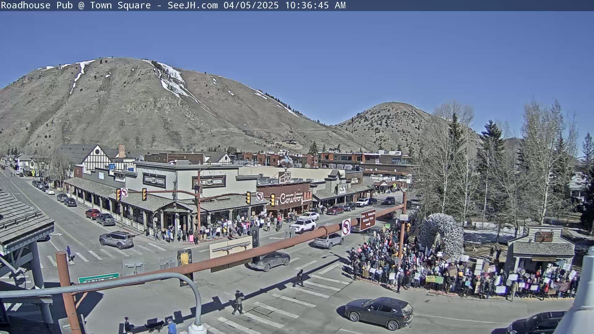A large crowd gathers in a town square, situated beneath mountains on a sunny day, with cars driving on the street nearby.