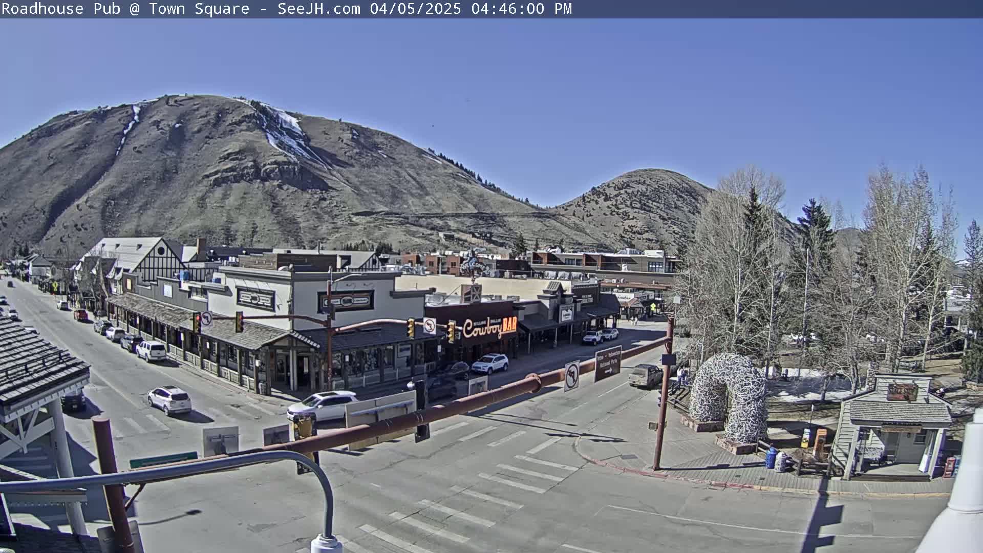 A sunny day reveals a mountain town square with buildings, cars, and trees, nestled between two hillsides.