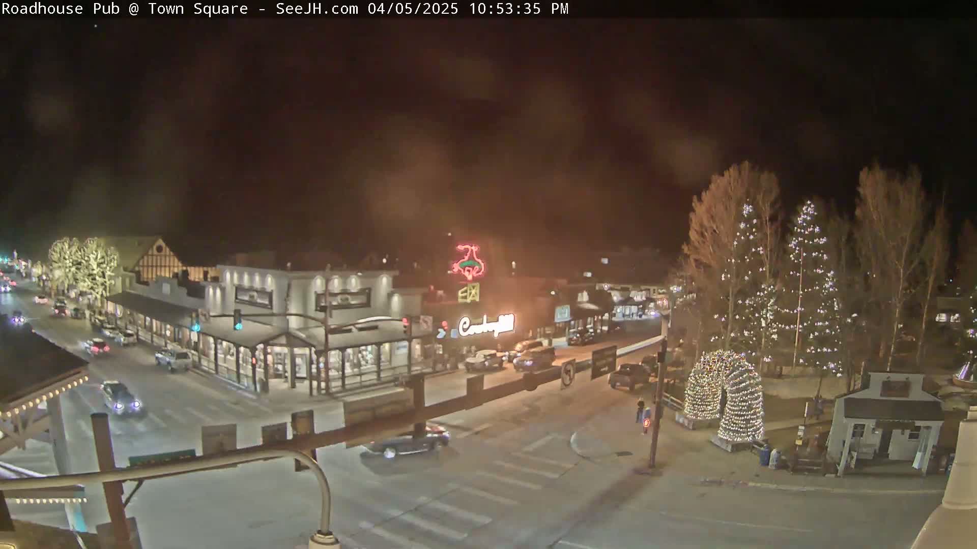A nighttime view of a town square with several buildings, cars, and Christmas lights illuminating the area under a hazy sky.