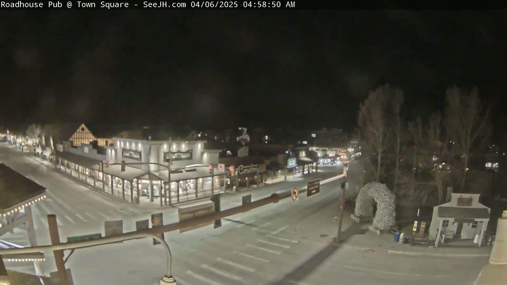 A snow-covered town square at night, with buildings lit and a few vehicles on the road.