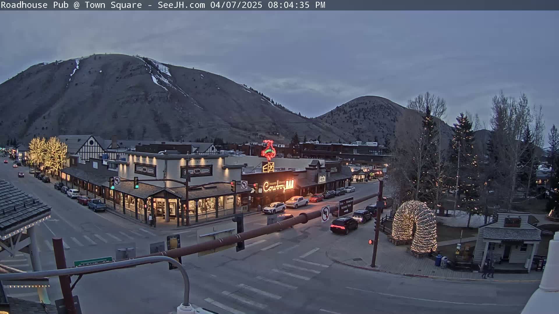 A town square at dusk, under a twilight sky, with cars parked along the streets and buildings decorated with holiday lights.