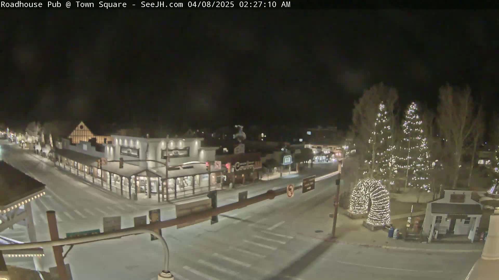 A snow-covered town square at night, illuminated by streetlights and Christmas lights strung on trees and a decorative arch.