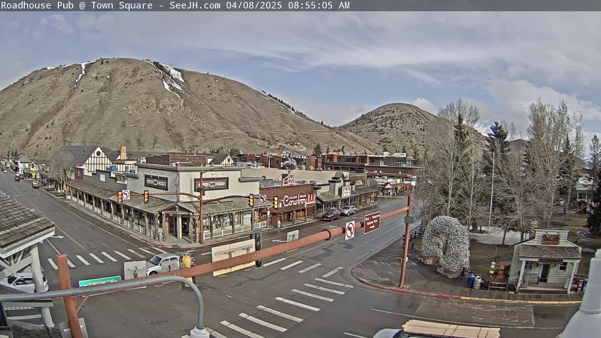 A partly cloudy day reveals a town square nestled between two hills, with several buildings lining a street, and sparse traffic.