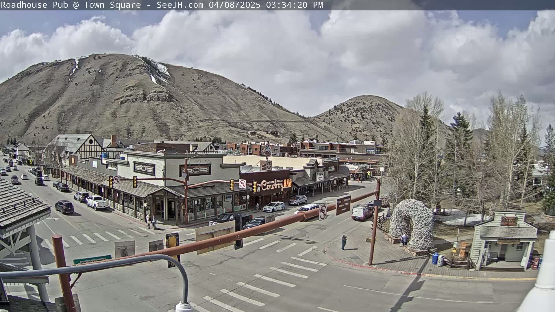 A partly cloudy day reveals a mountain town square with buildings lining a street, cars parked along the sides, and a few people walking around.