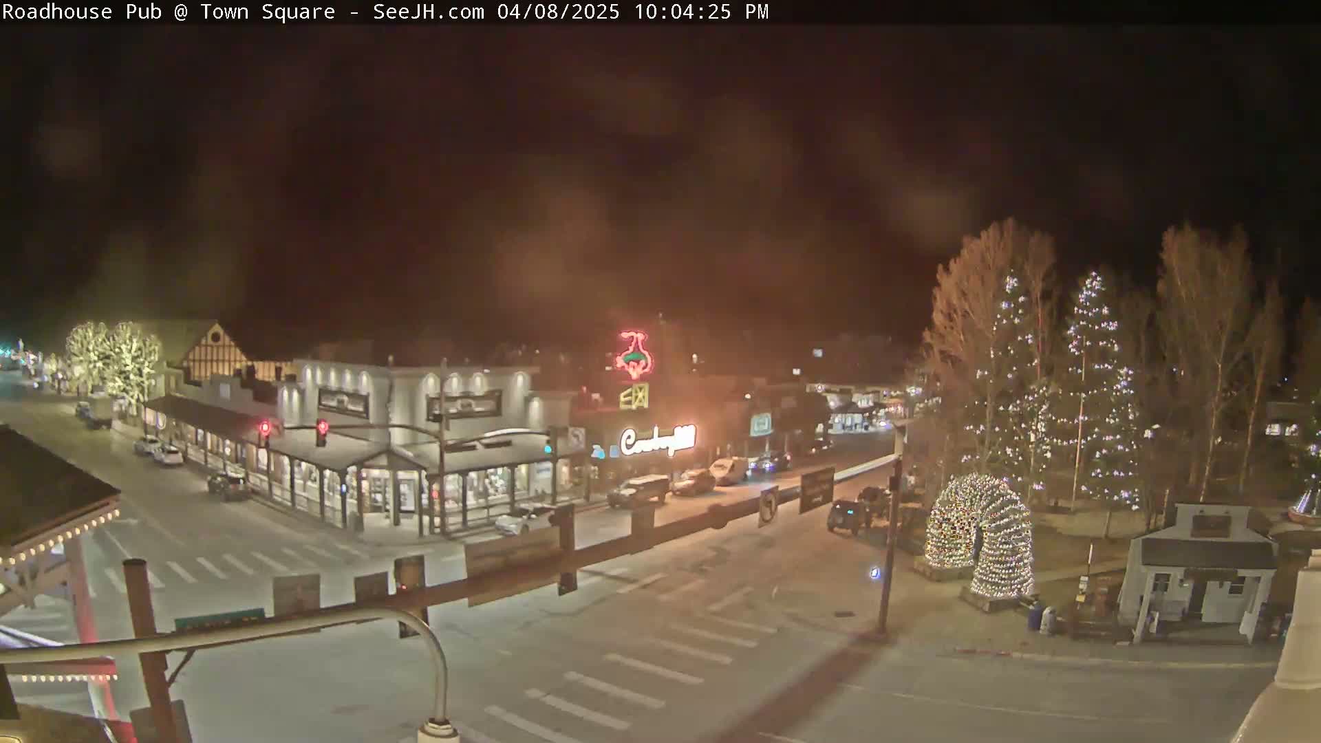 A nighttime, snow-covered town square features several illuminated buildings and Christmas trees.