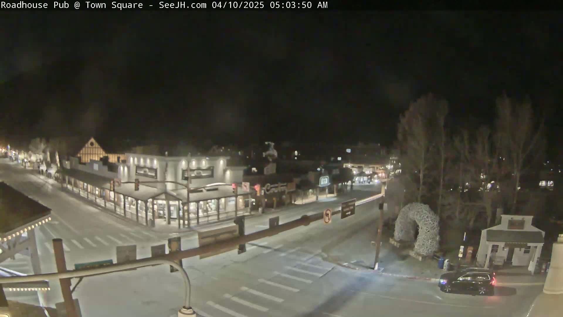 A nighttime view of a snow-covered town square with several buildings and a single car parked near a small structure.