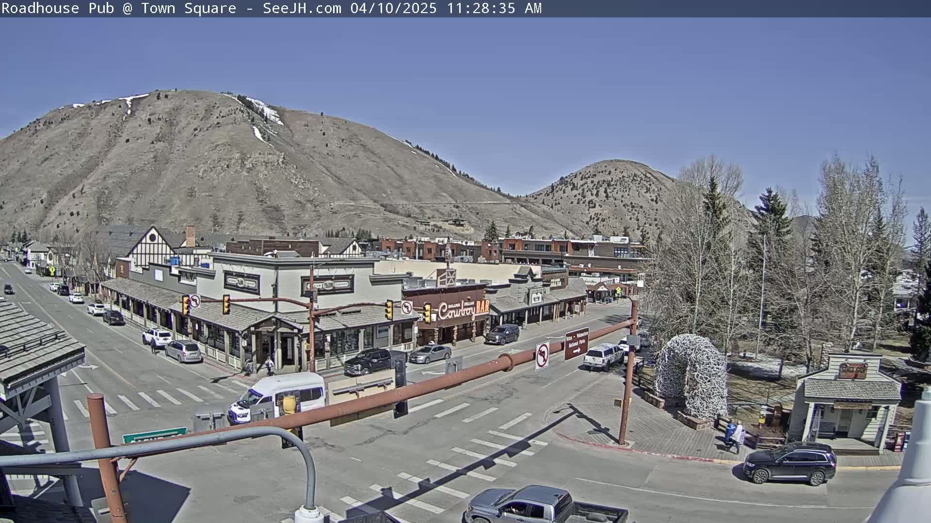 A sunny day reveals a town square with buildings, vehicles, and people, nestled beneath a large, barren mountain.