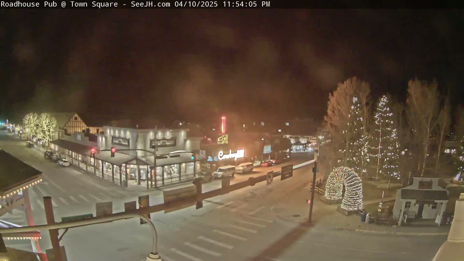 A nighttime, snowy view of a town square with illuminated buildings and Christmas lights on trees.