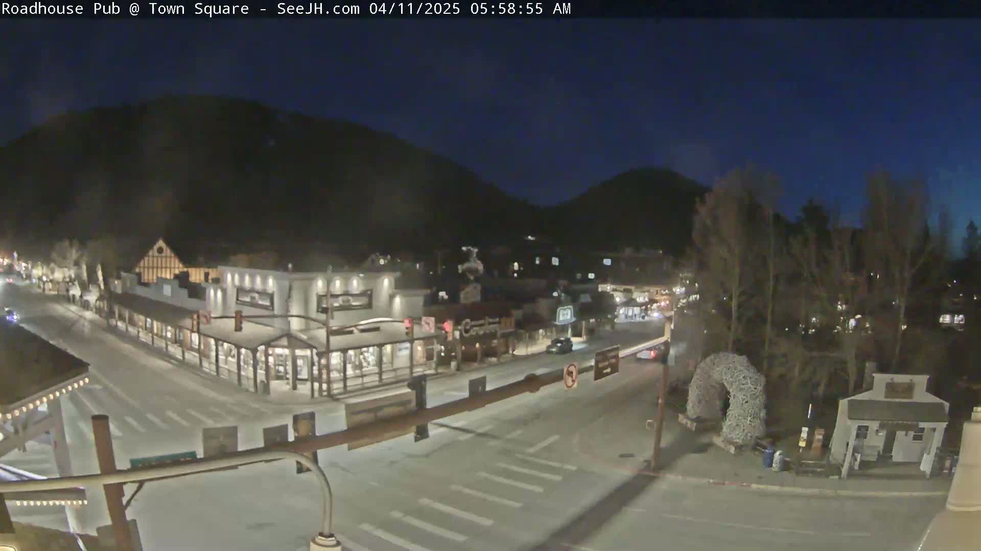 A nighttime view of a town square with several buildings, a few cars, and dark mountains in the background under a clear, dark sky.