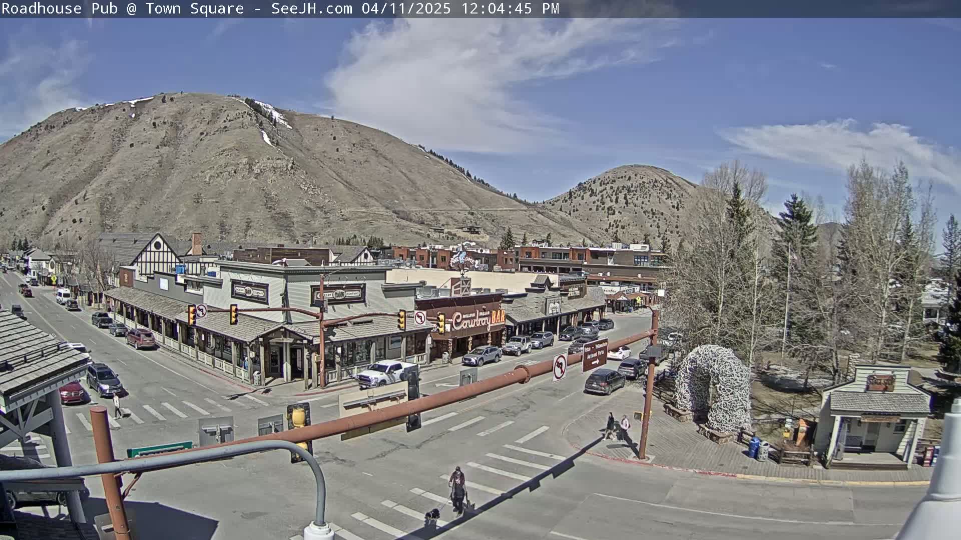 A sunny day in a mountain town shows a main street with buildings, cars, and pedestrians, with mountains in the background.