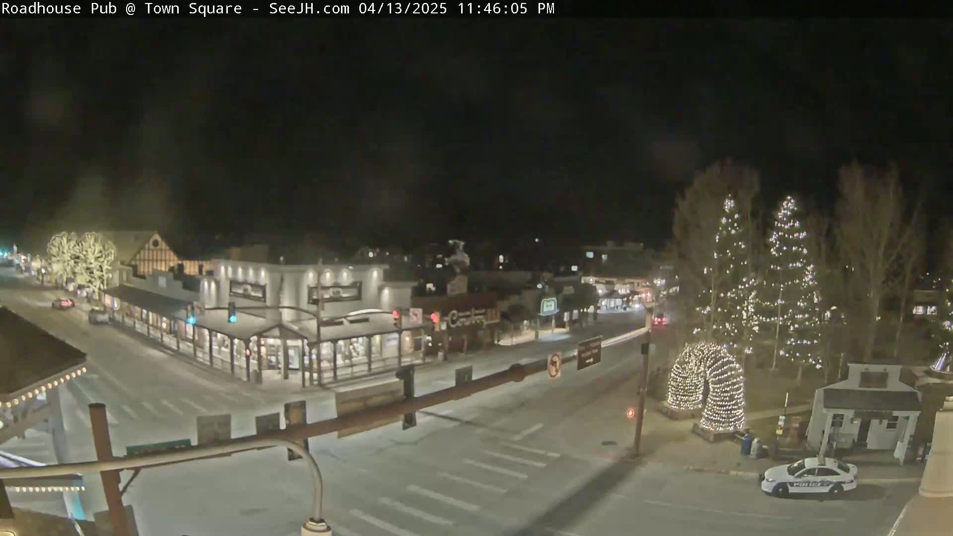 A nighttime, snow-covered town square is viewed from above, showing several buildings, illuminated streets, and decorated trees.