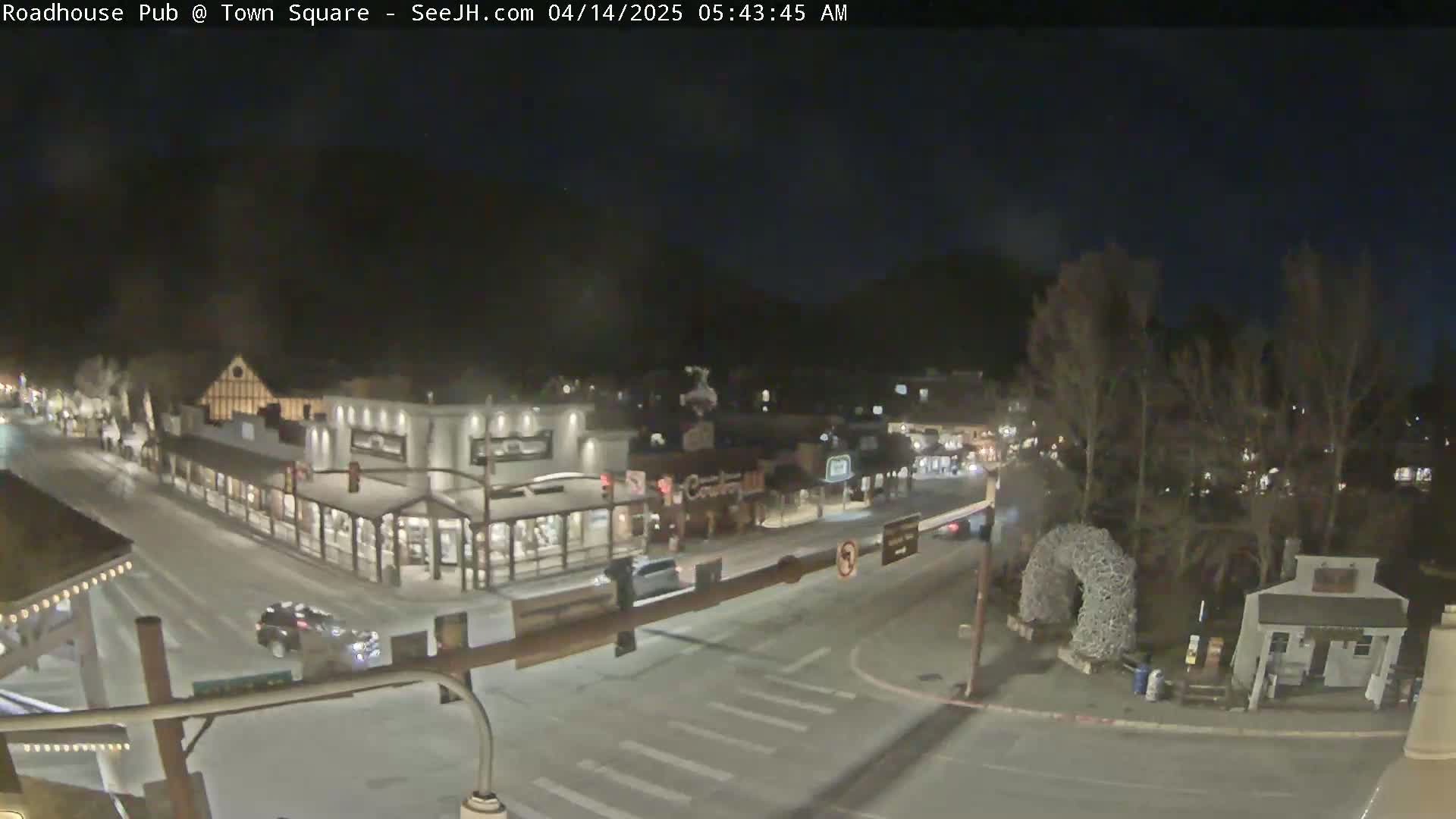 A snow-covered town square at night, viewed from above, shows several buildings with illuminated storefronts and a few vehicles on the street.