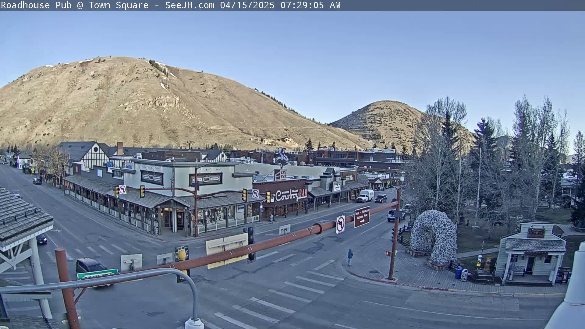 A mostly clear, sunny day reveals a town square nestled between two hills, with several buildings, vehicles, and pedestrians visible.