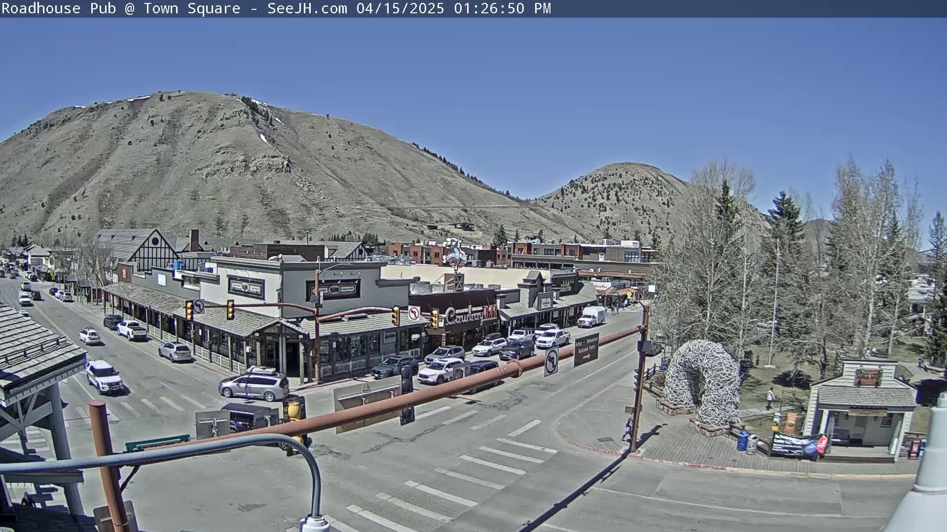 A sunny day in a town square, featuring a street with parked and moving cars, buildings lining the street, and a large hill in the background.