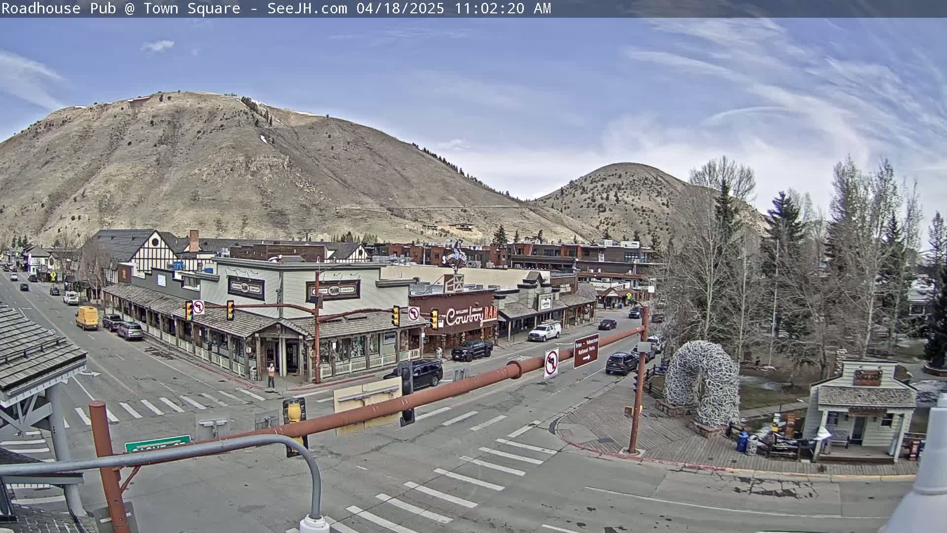 A mostly sunny day reveals a town square nestled between two hills, with several buildings lining the streets and sparse traffic.