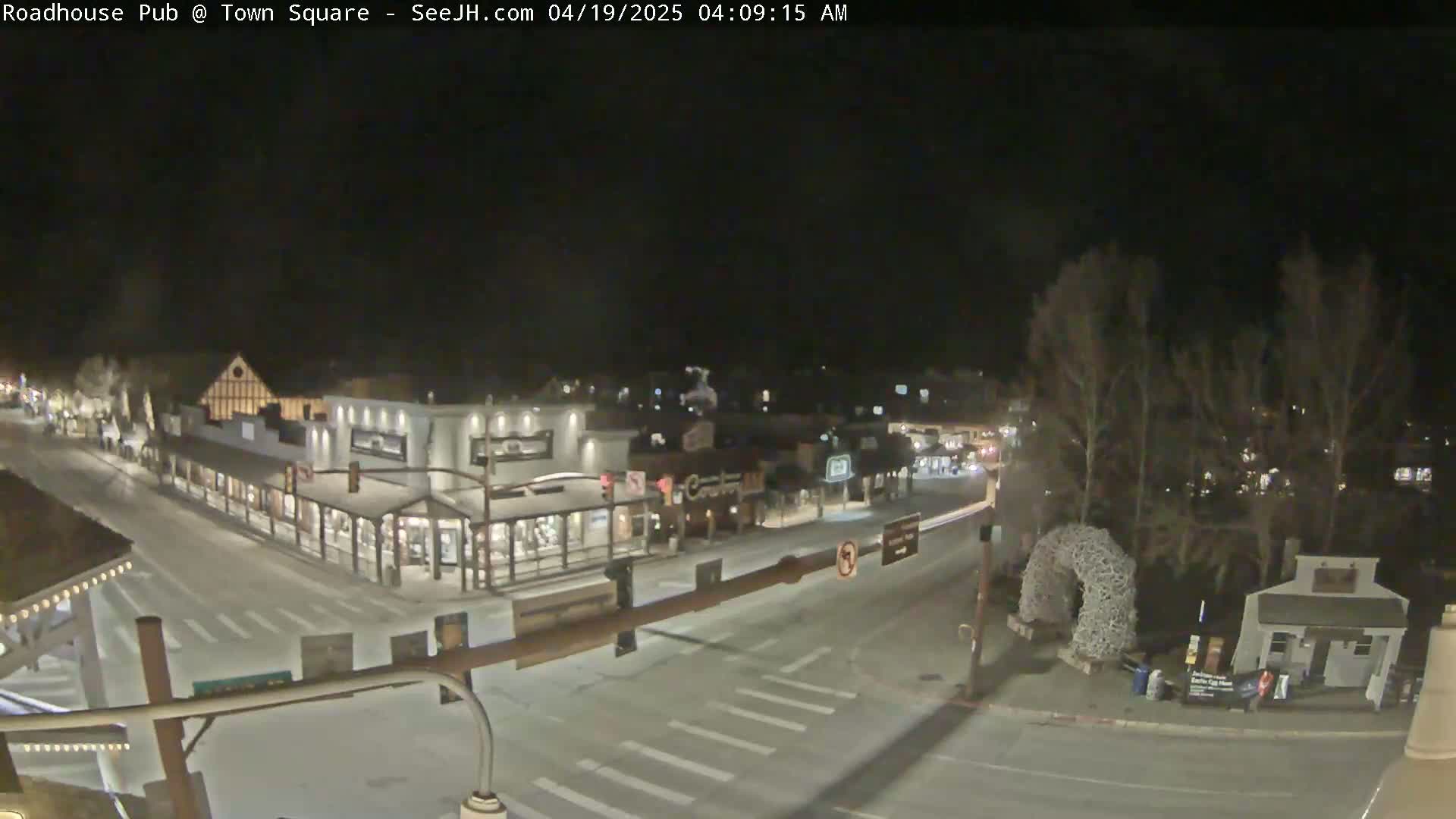 A snow-covered town square at night, illuminated by streetlights, shows several buildings, a street intersection, and sparse vehicle traffic.