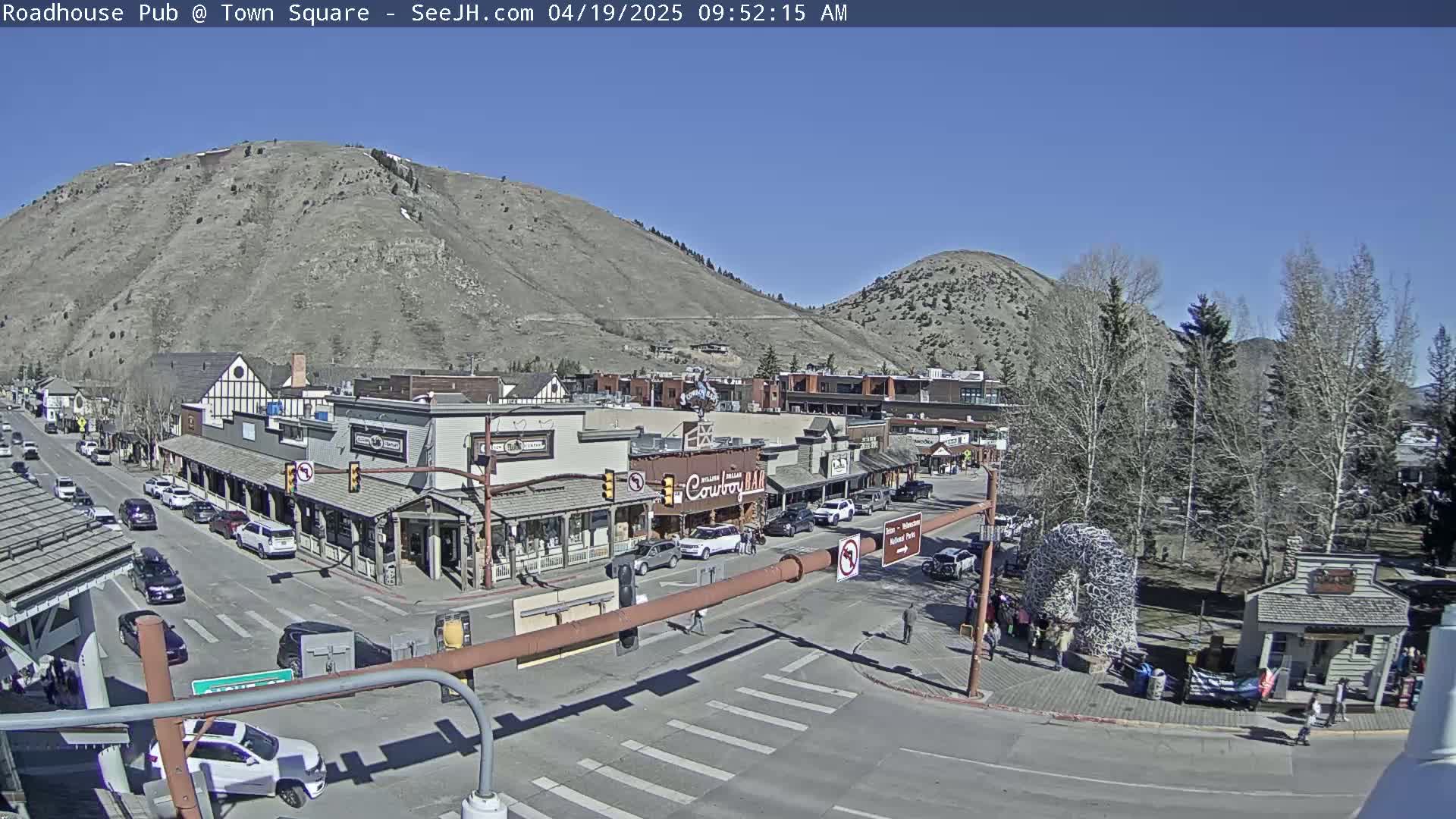 A sunny day reveals a town square nestled at the base of a mountain, with cars parked along the streets and people walking around.